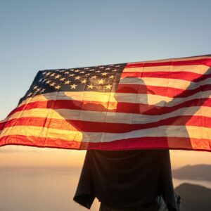 Proud man raising the flag of the United States of America standing on the top of the mountain at sunset